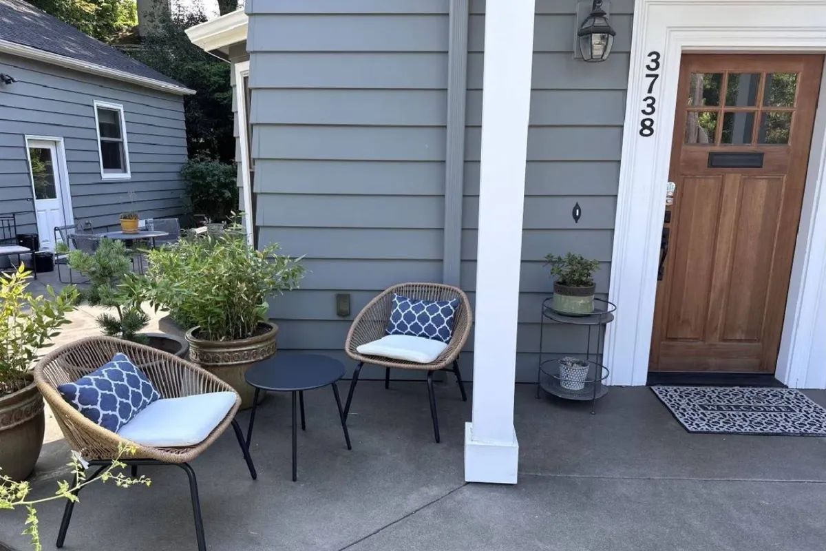 An inviting exterior scene featuring a comfortable seating area by the front door, surrounded by lush planters. The setting suggests relaxing outdoors, sipping morning coffee, or enjoying quiet conversations in the fresh air.