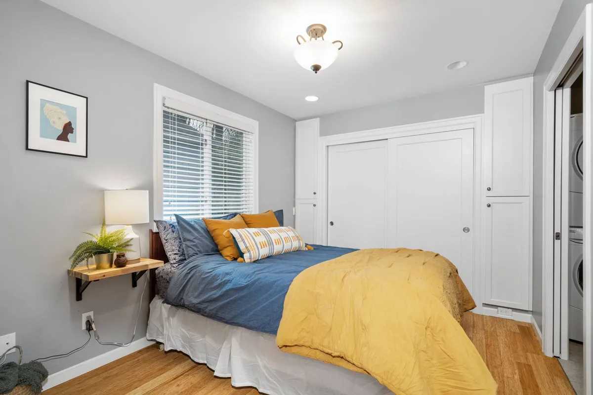 A comfortable bedroom with a queen bed adorned in yellow and blue bedding, accentuated by framed local artwork and a bedside lamp. Sunlight filters through the blinds, creating a peaceful and warm ambience perfect for restful sleep or morning inspiration.
