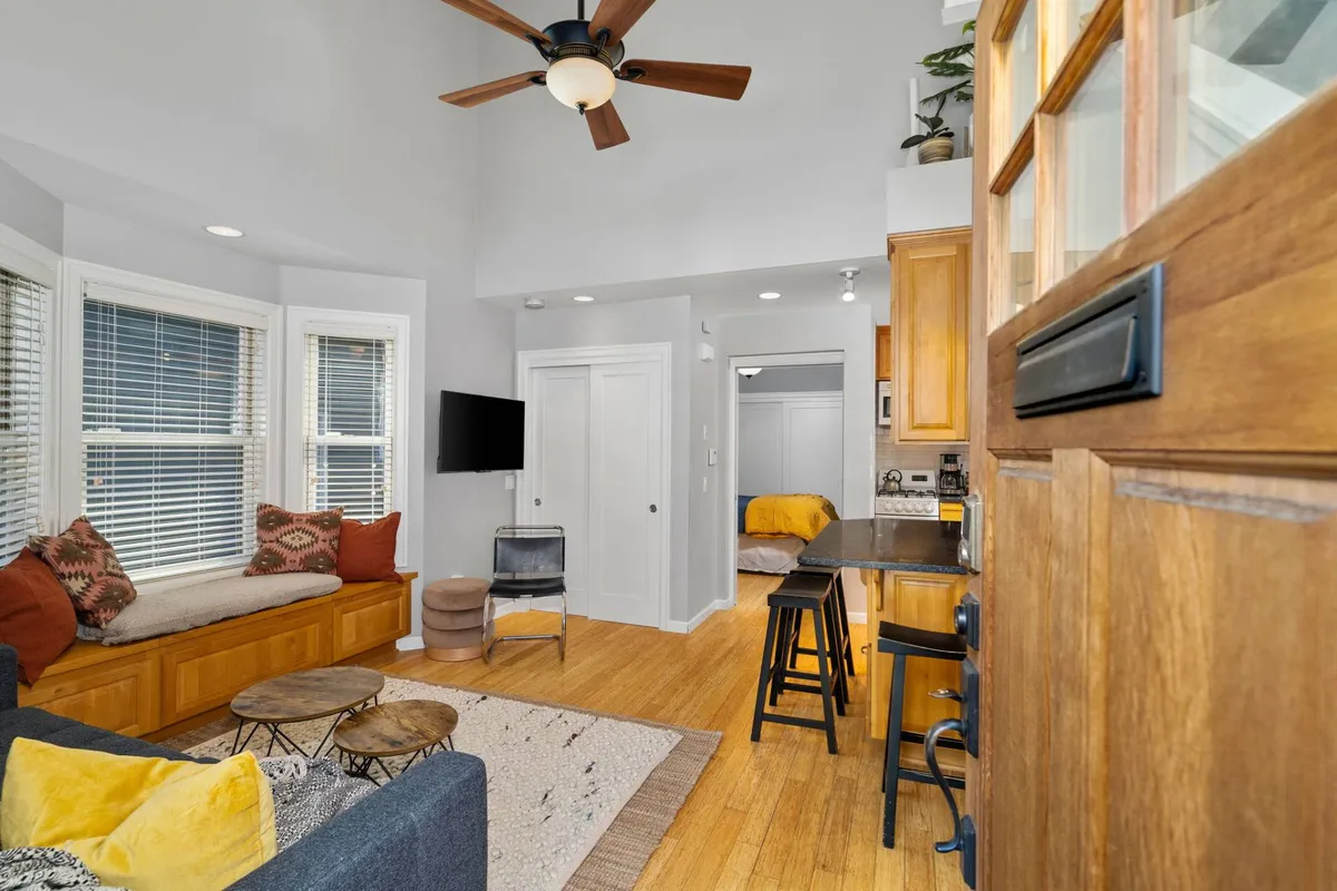 A modern, comfortable seating area is paired with a kitchen bar in an open-plan space. Large windows flood the room with natural light, and a bench near the window offers a quiet nook for reading or brainstorming. The overall mood is both cozy and vibrant, promoting both relaxation and social interaction.