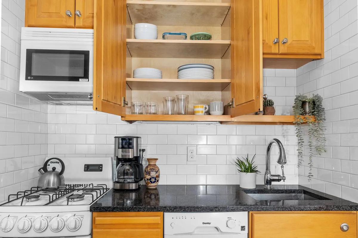 A kitchen nook with a sleek black granite countertop, open cabinet displaying dishware, a coffee maker, and essential kitchen appliances. The space is warm and homey, designed for shared meal preparation, casual breakfasts, or hosting friends over coffee.