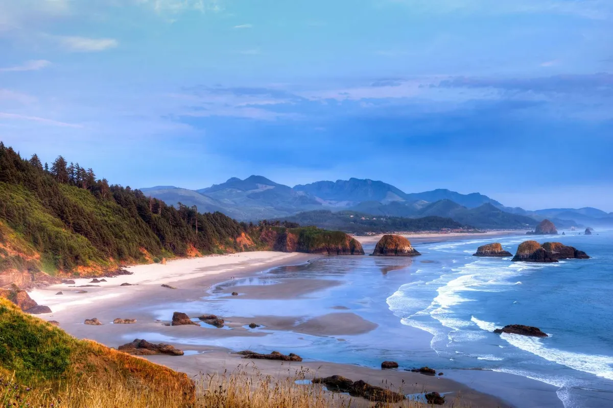 A stunning view of a tranquil beach scene, featuring sweeping ocean waves, rugged rock formations, distant mountains, and a fringe of lush forest. The scene is open, inviting, and evokes a sense of adventure and natural beauty—perfect for visitors who want to explore the region's outdoor attractions.