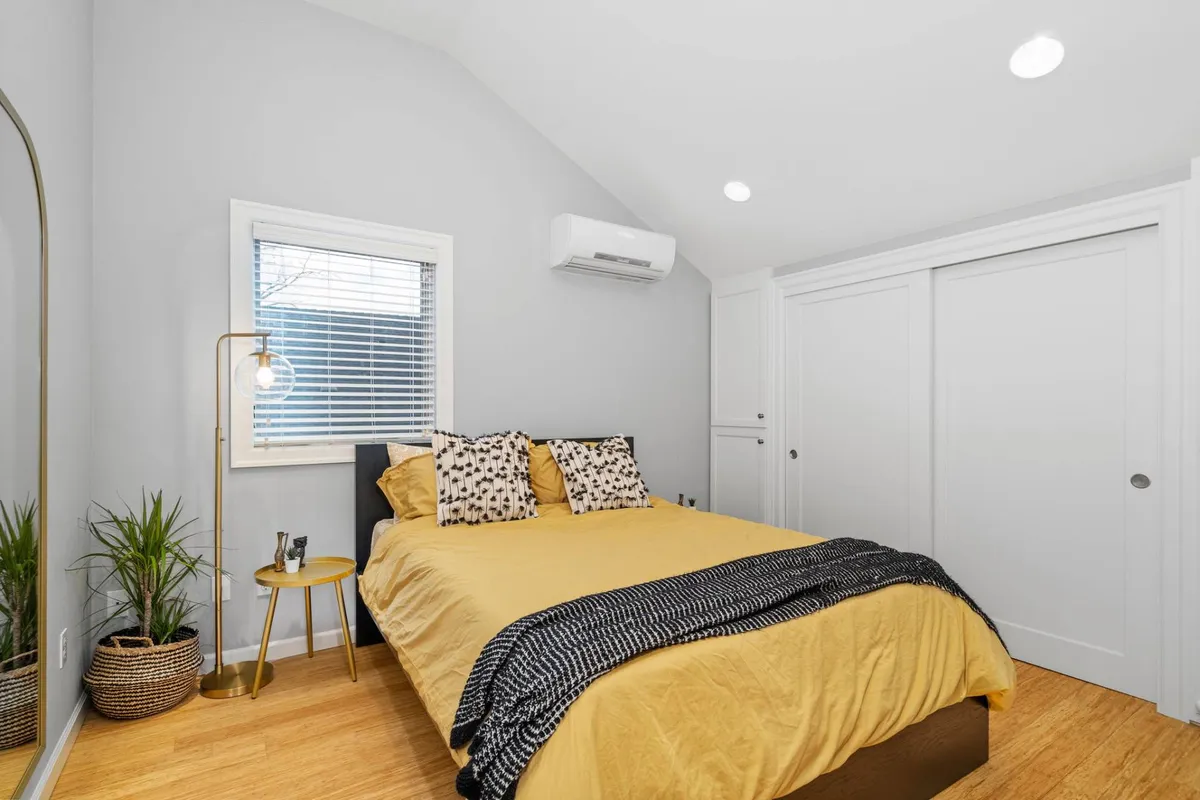 A tranquil bedroom featuring a queen bed dressed with quality linens and decorative pillows. Sunlight streams in through the window, highlighting a cozy reading nook and lush green plants that add freshness to the space. The overall mood is inviting and restful, ideal for unwinding after a long day.