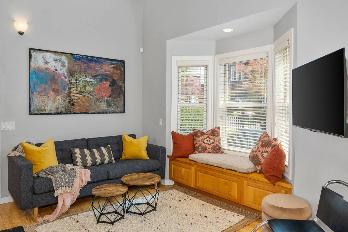 An interior view of a living area with a comfortable sofa, a spacious bay window that lets in abundant light, coffee tables for gathering, and distinctive artwork adorning the wall. The composition suggests a perfect blend of relaxation, conversation, and art appreciation.