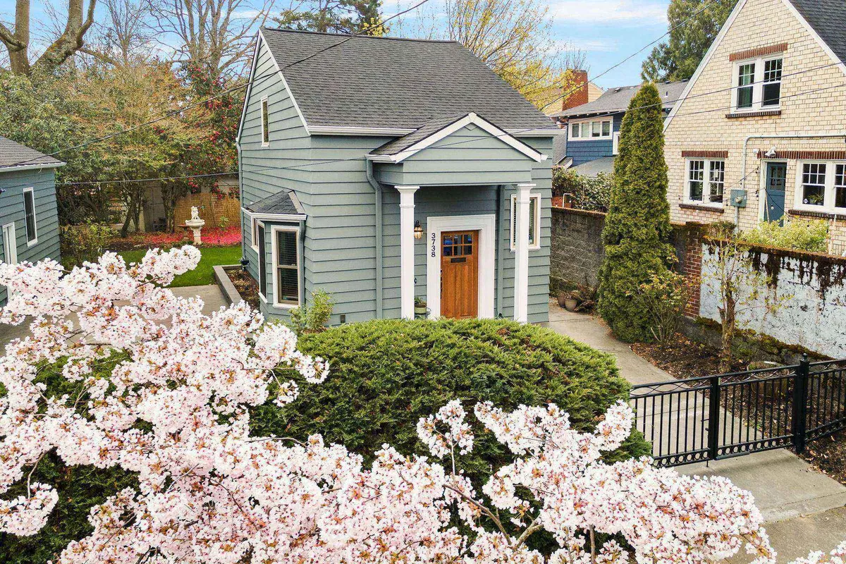 The image showcases the exterior of a charming blue-gray single-family house framed by a blooming cherry blossom tree. The welcoming entrance door and inviting front yard set a cozy, springtime scene, with glimpses of neighboring houses that suggest a friendly, vibrant neighborhood.