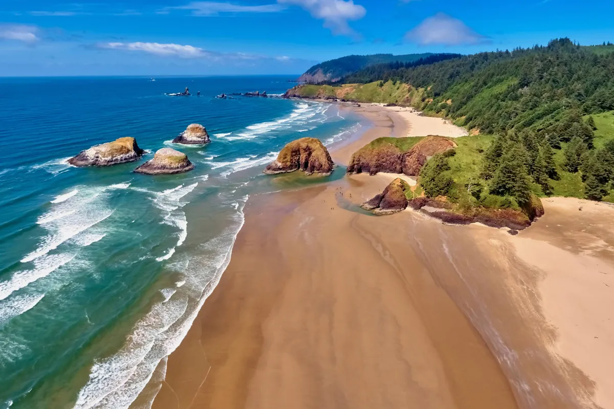 A serene beach scene with dramatic rock formations framed by waves, soft sand, and lush greenery. The image exudes a peaceful, exploratory mood, suggesting opportunities for photography, tide pool exploration, and relaxation by the water.